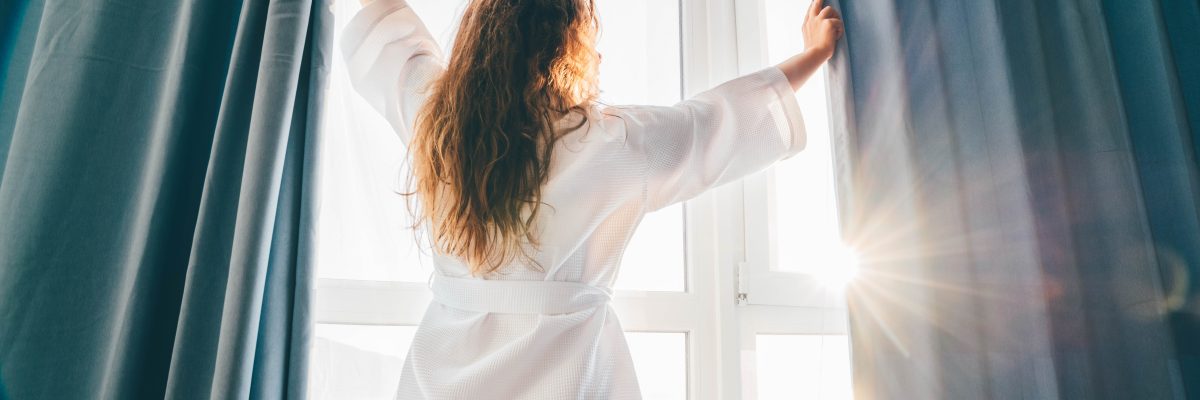 Woman opening curtains in room at sunrise.